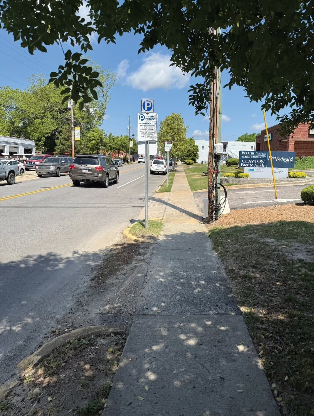 A public parking sign with a blue “P” symbol and several lines of small rules stands beside a downtown Clayton sidewalk near a bank parking lot. Cars travel and park along the street.
