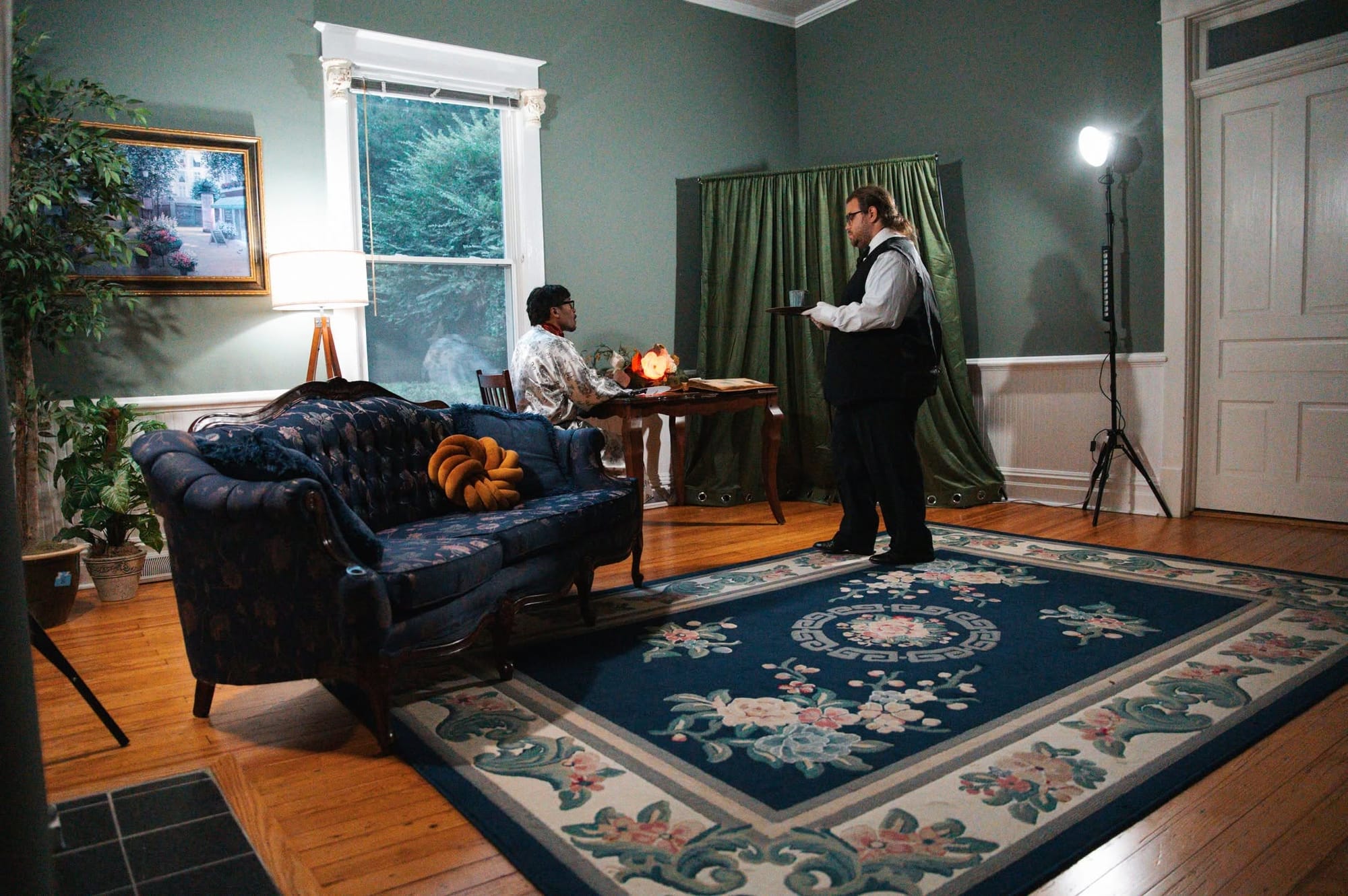 Two performers act out a scene in a furnished room, with one seated at a small table and another standing nearby holding a tray.