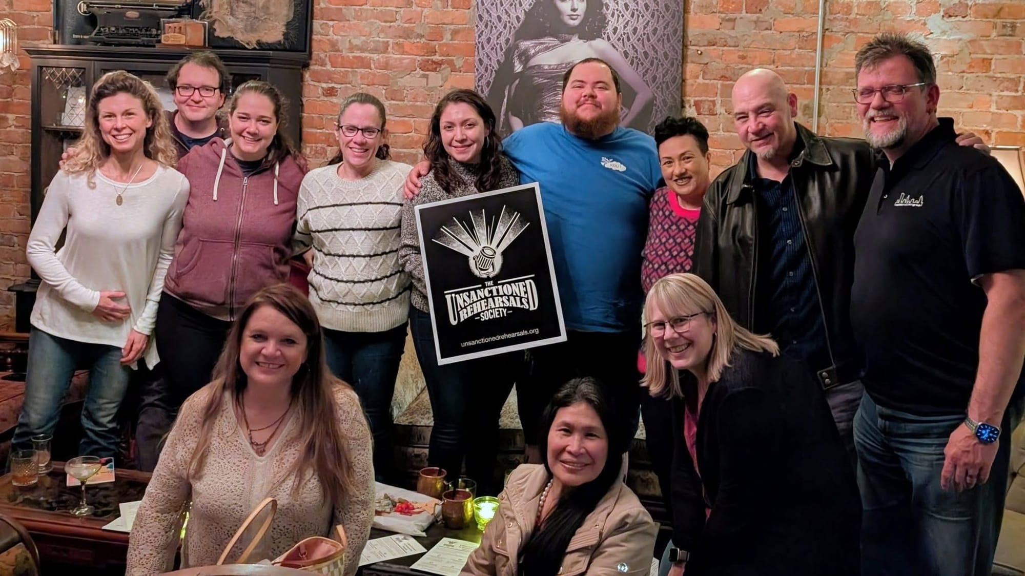 A group of Unsanctioned Rehearsal Society members pose together indoors around a black-and-white sign bearing the theater group’s logo.