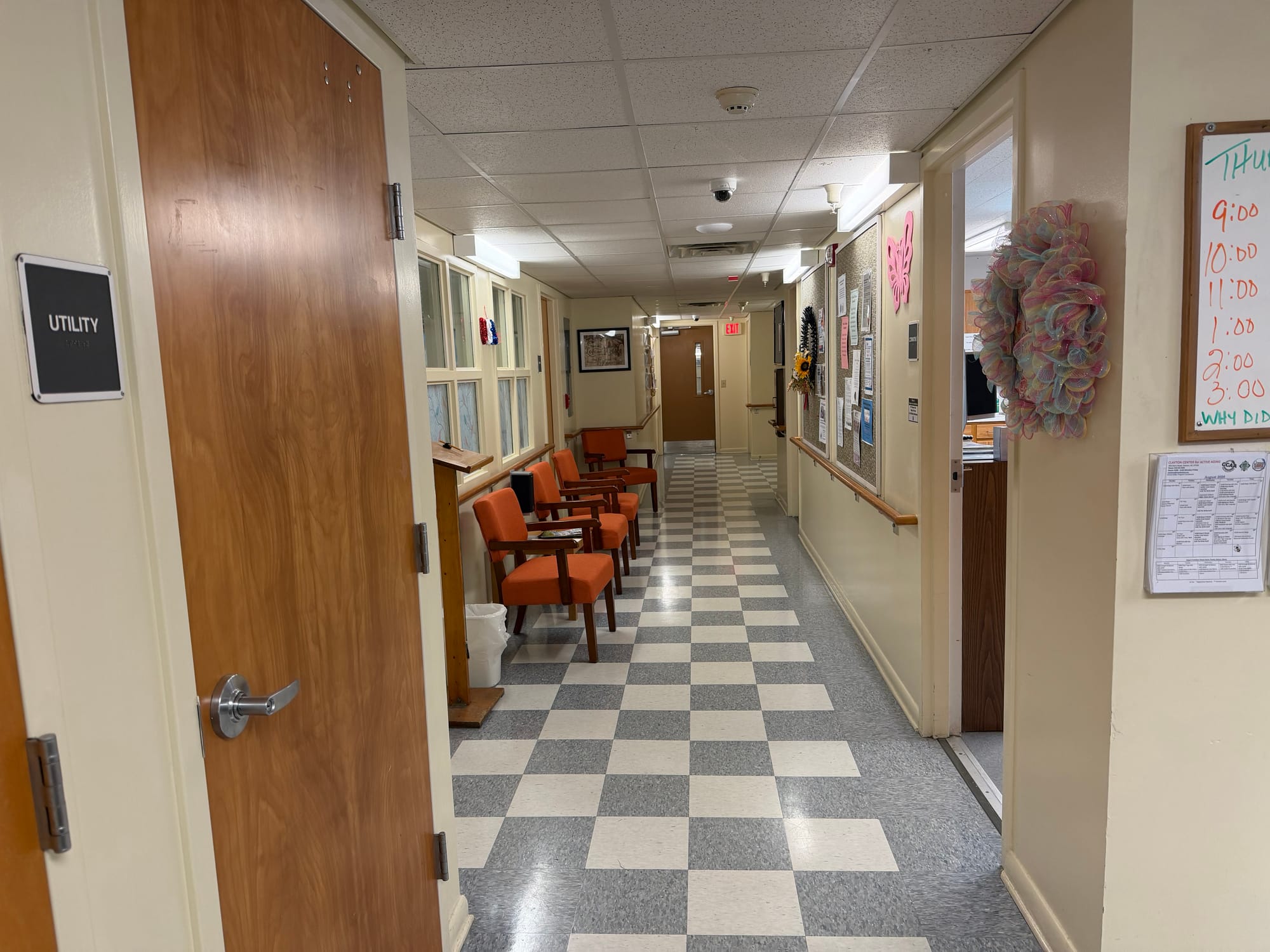 A narrow hallway inside the Clayton Center for Active Aging with orange chairs lining one wall, bulletin boards, office doors and a black-and-white checkered floor.