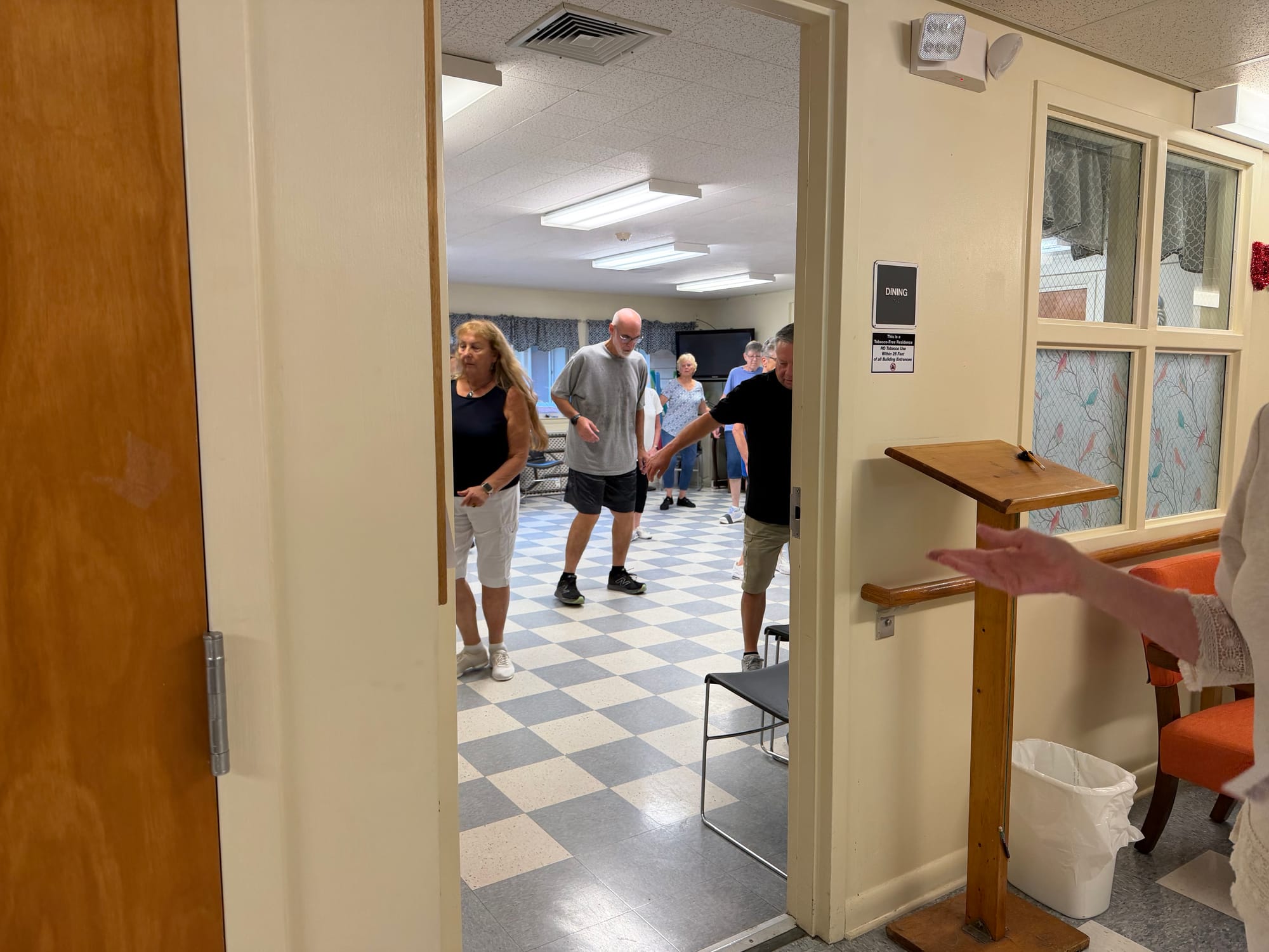 A group of adults participating in an exercise class inside the Clayton Center for Active Aging, viewed through the doorway of a room marked “Dining.”