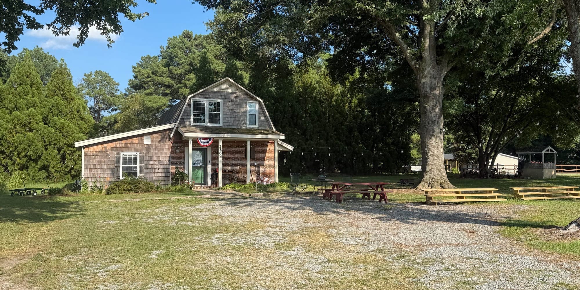 The Barn House, a small historic barn-style building on Barber Mill Road in Clayton, sits beside a large shade tree with picnic tables and a gravel area out front.