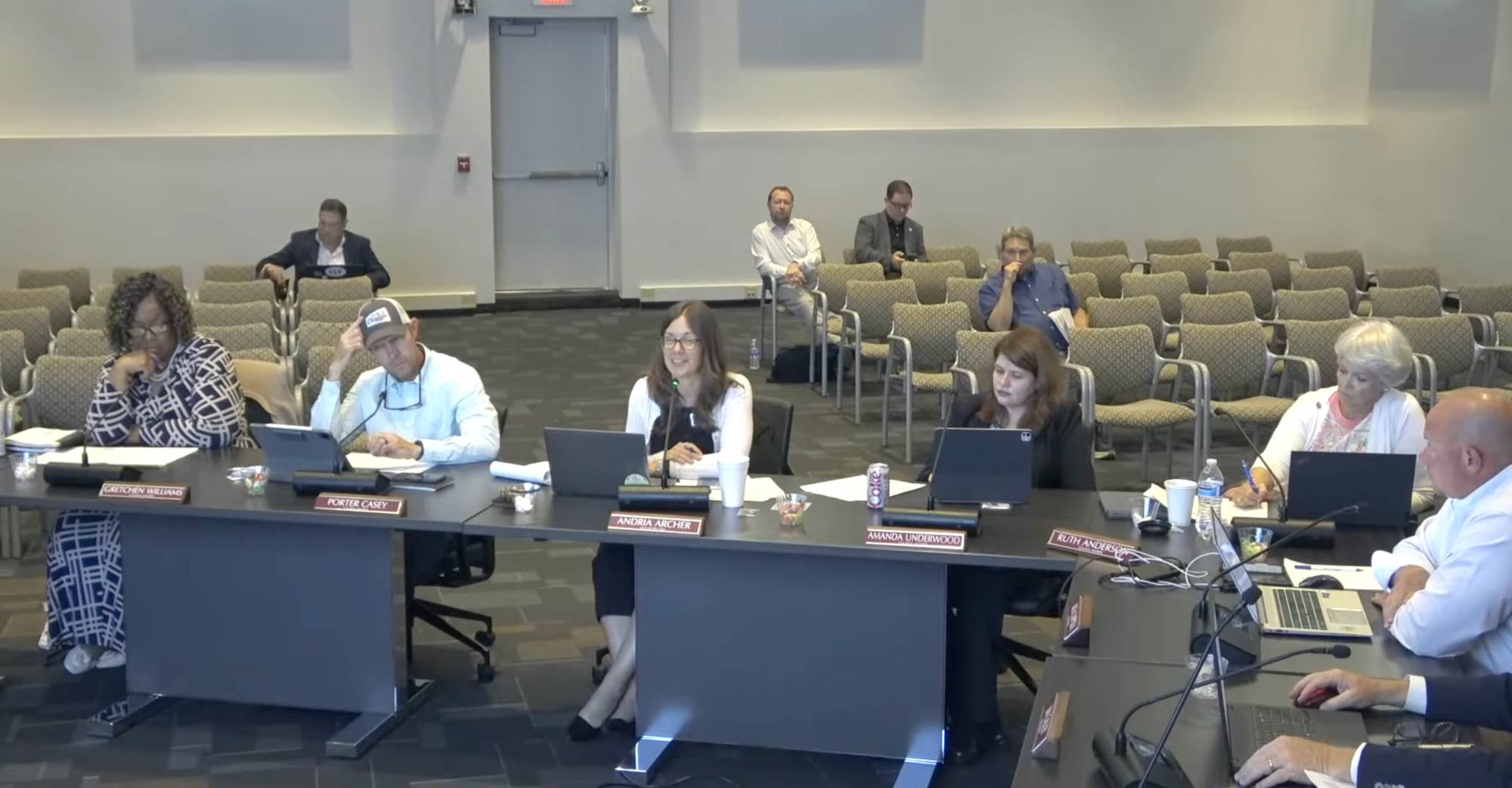 Five Clayton Town Council members sit behind desks with laptops and meeting materials as they discuss downtown parking during a work session. Several staff members and attendees sit in the mostly empty council chamber behind them.
