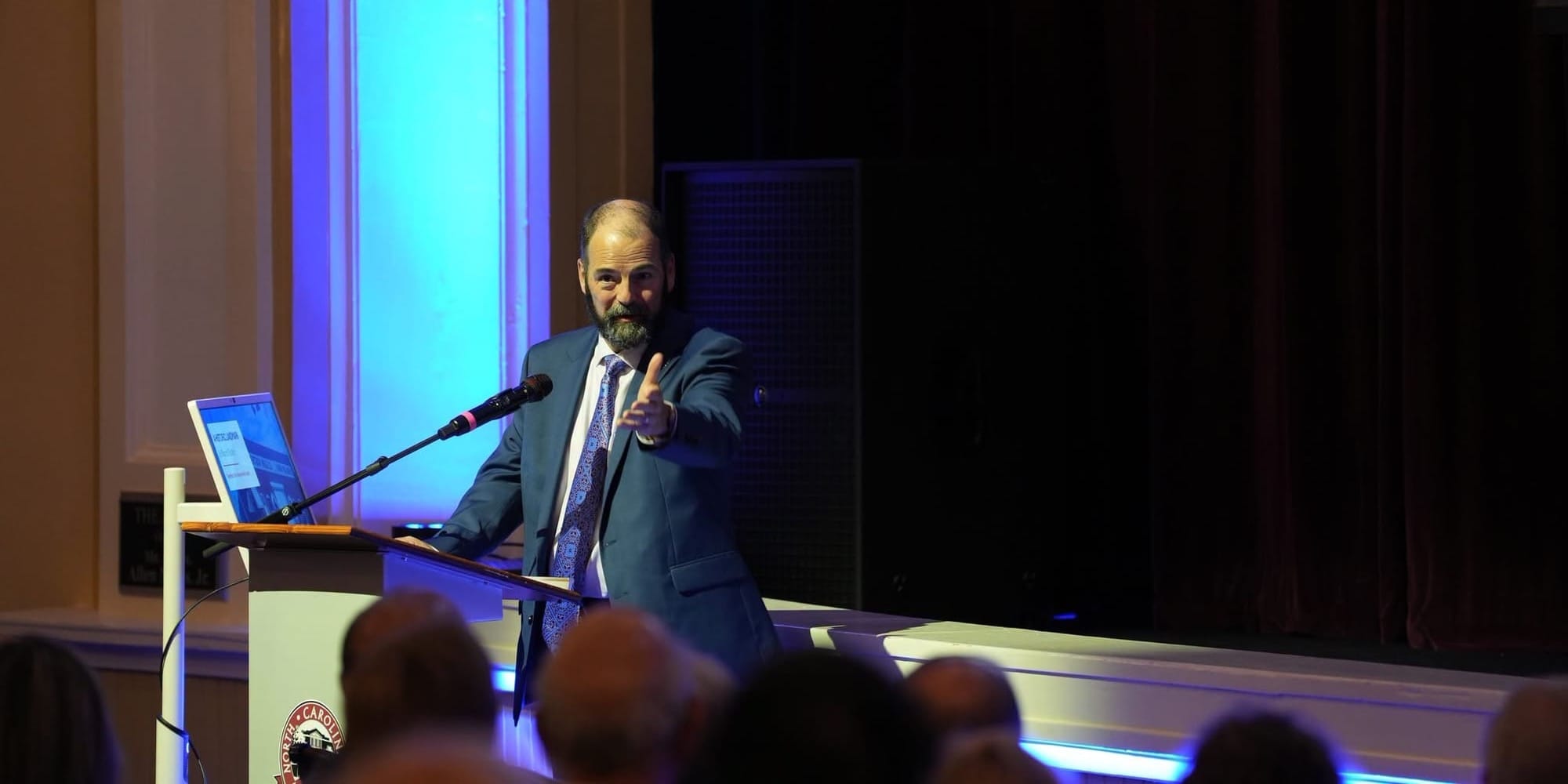 Chappell Jordan of Jacobs stands at a podium speaking to an audience during a Town of Clayton presentation. He is wearing a dark suit and gesturing with one hand as blue stage lighting illuminates the wall behind him.