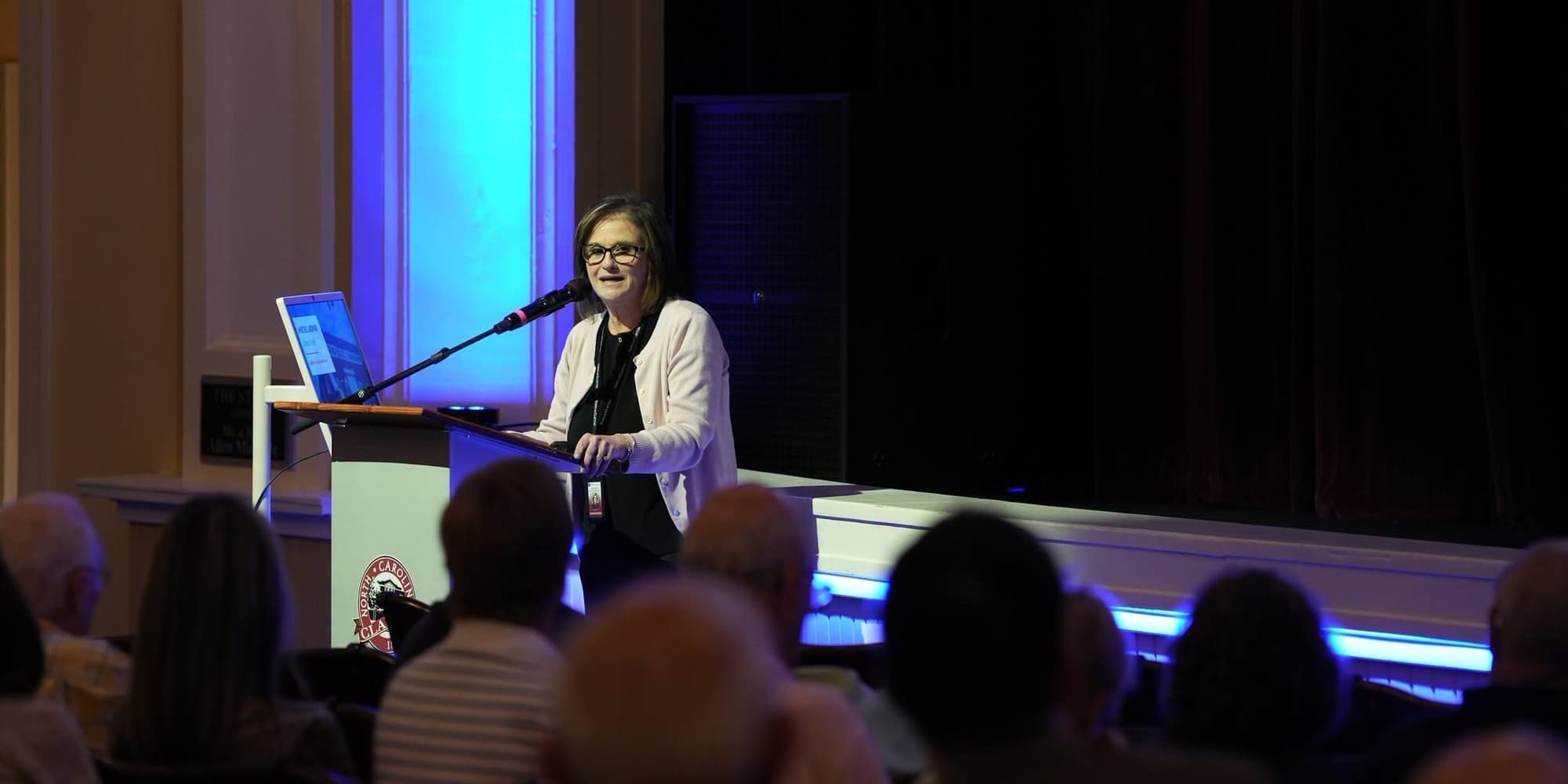Deputy Town Manager Dolores Gill stands at a podium speaking to an audience inside The Clayton Center during a Town presentation. Blue stage lighting illuminates the wall behind her.