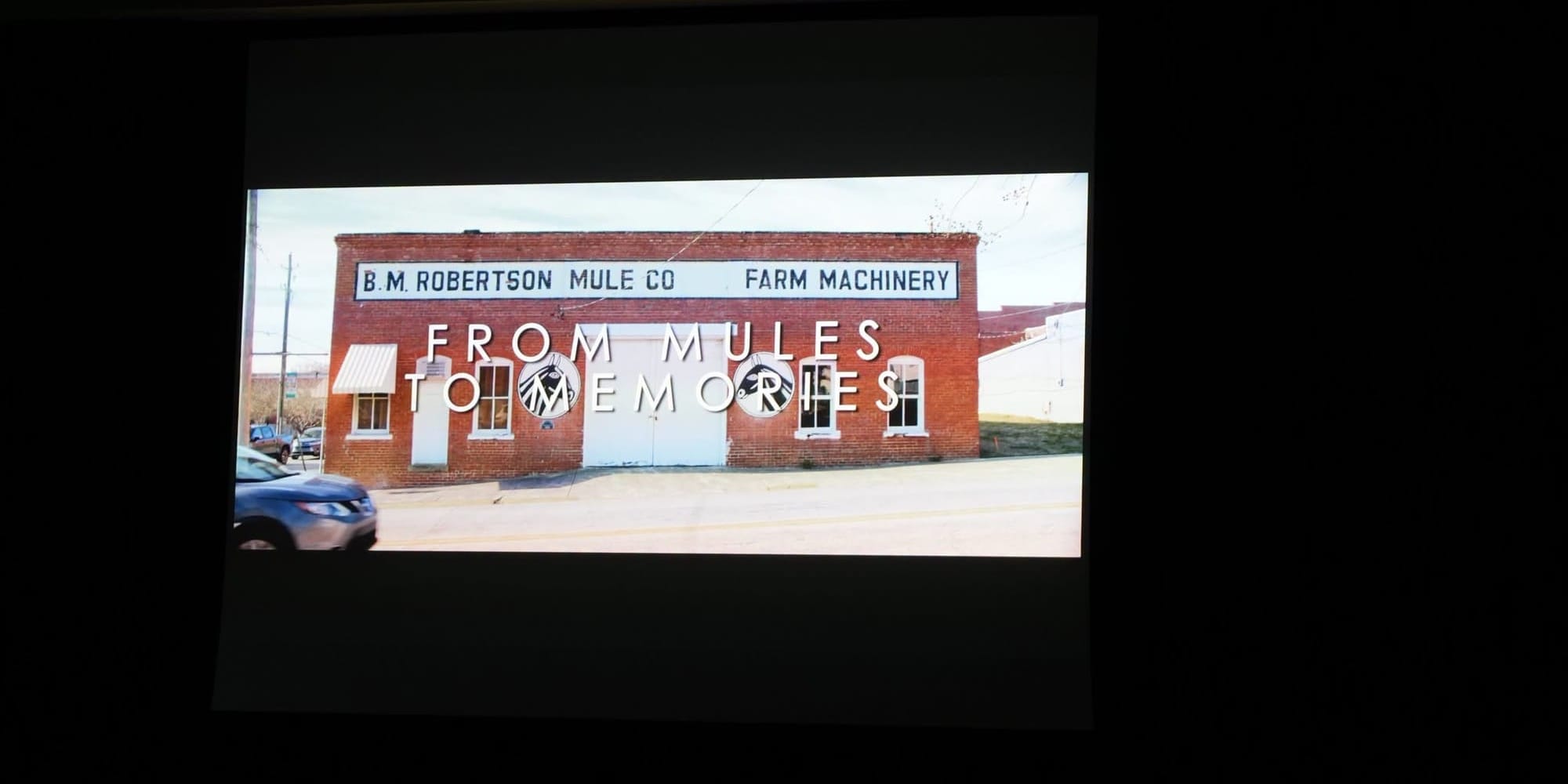 A projection screen inside The Clayton Center shows the title card for From Mules to Memories, featuring a photograph of the historic B.M. Robertson Mule Company building in Downtown Clayton.