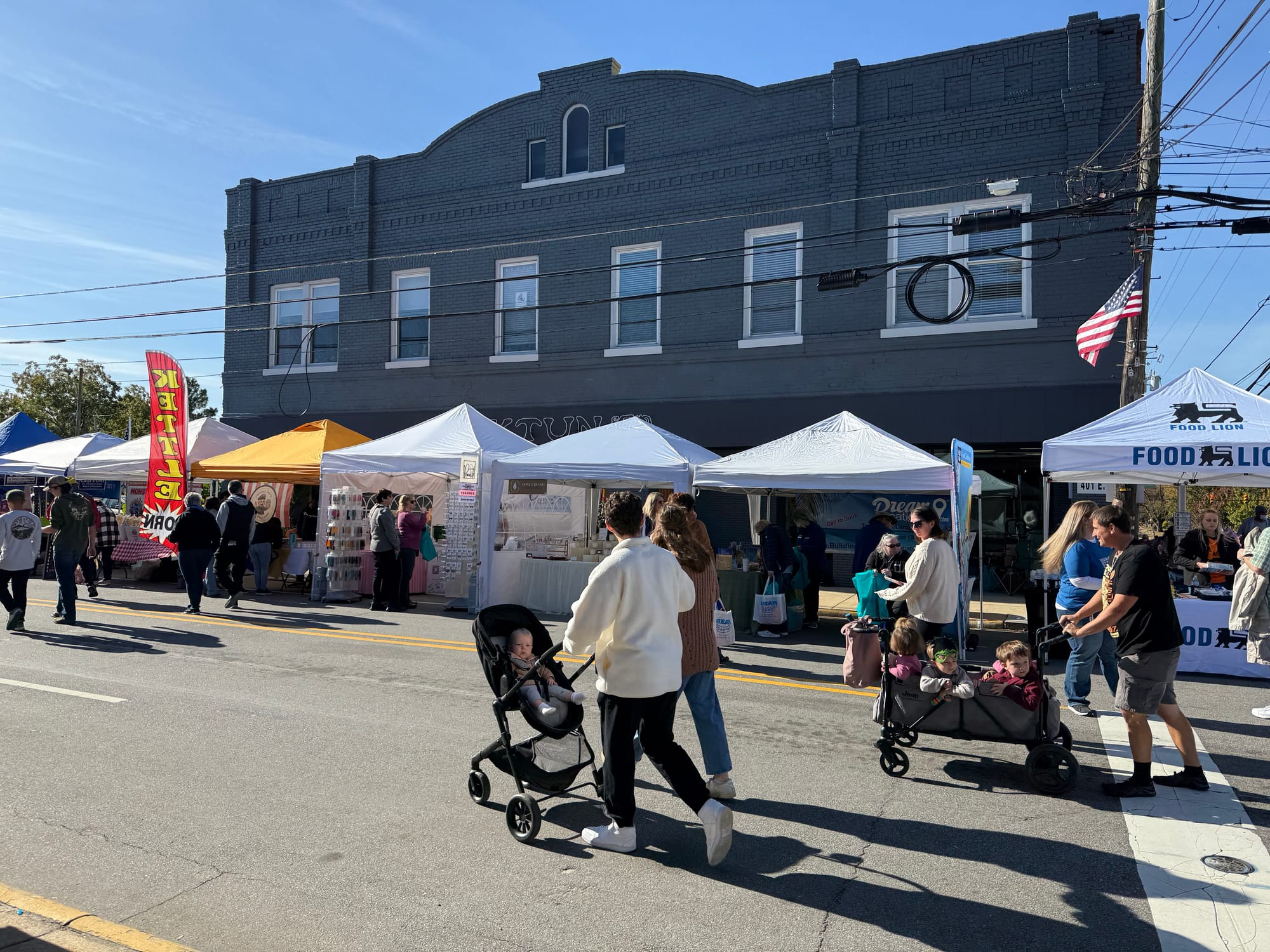 A row of white and colorful vendor tents lines the street in front of a large, dark brick commercial building housing Aaktun in downtown Clayton. Festivalgoers walk along Main Street, including families pushing strollers and wagons, while the tents partially obscure the businesses behind them.