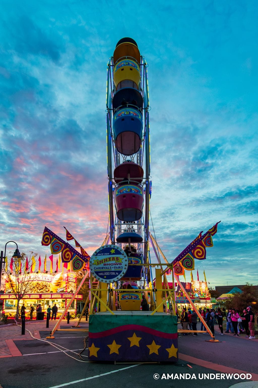 A colorful Ferris wheel photographed from directly in front at the Clayton Harvest Festival. Brightly lit carnival food stands, decorative flags, and groups of festivalgoers surround the ride beneath a dramatic blue, pink, and orange sunset sky.