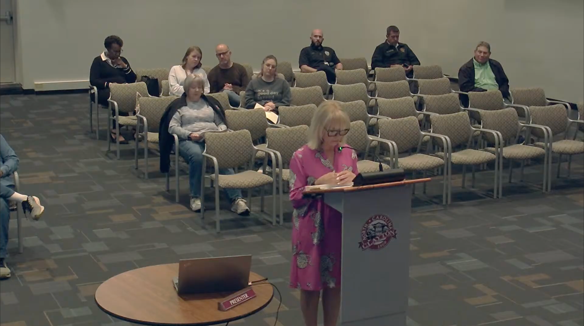 Dana Wooten stands at a podium bearing the Town of Clayton seal, wearing a pink floral dress and speaking into a microphone. Several attendees sit in rows of chairs behind her in the council meeting room.