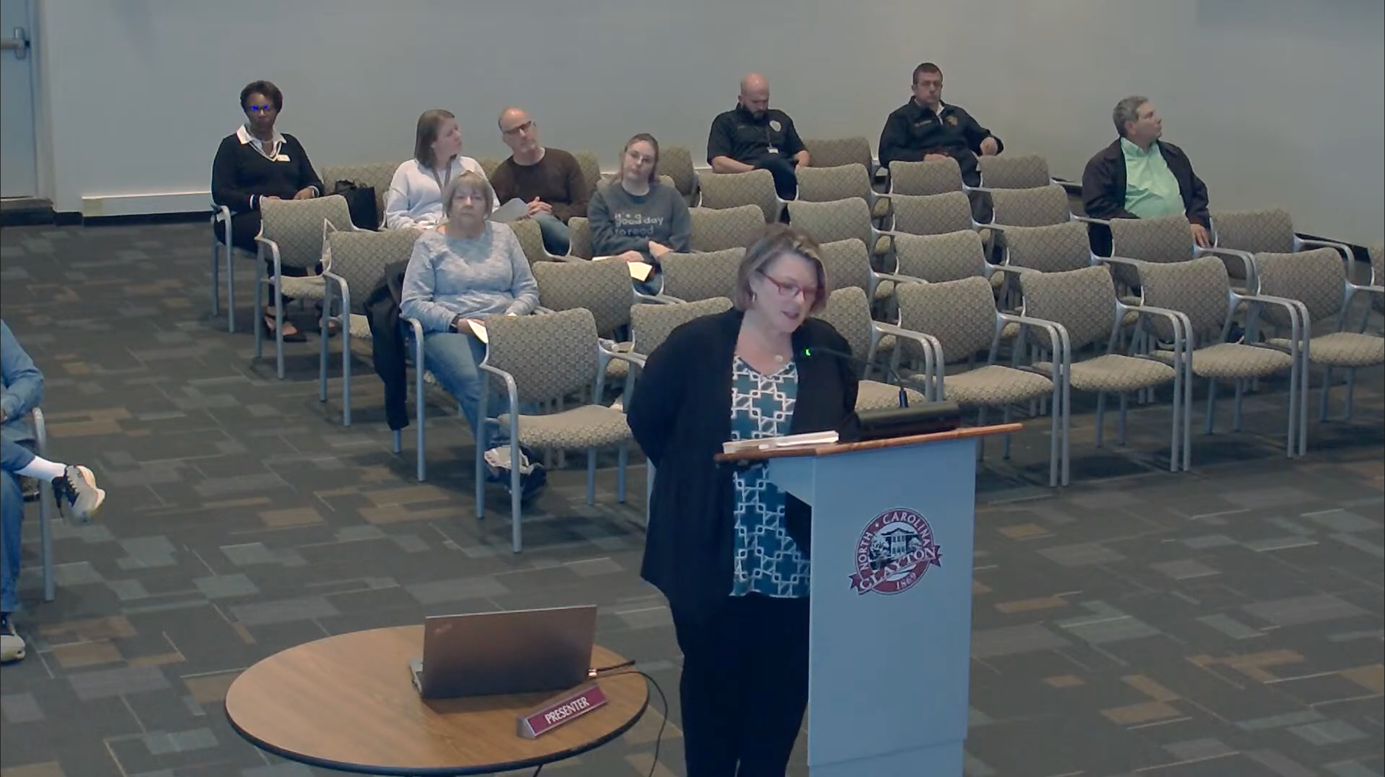 Melissa Overton stands at a podium bearing the Town of Clayton seal during a Town Council meeting. She wears glasses, a dark cardigan, and a blue-and-white patterned blouse. Several attendees sit in rows of chairs behind her.