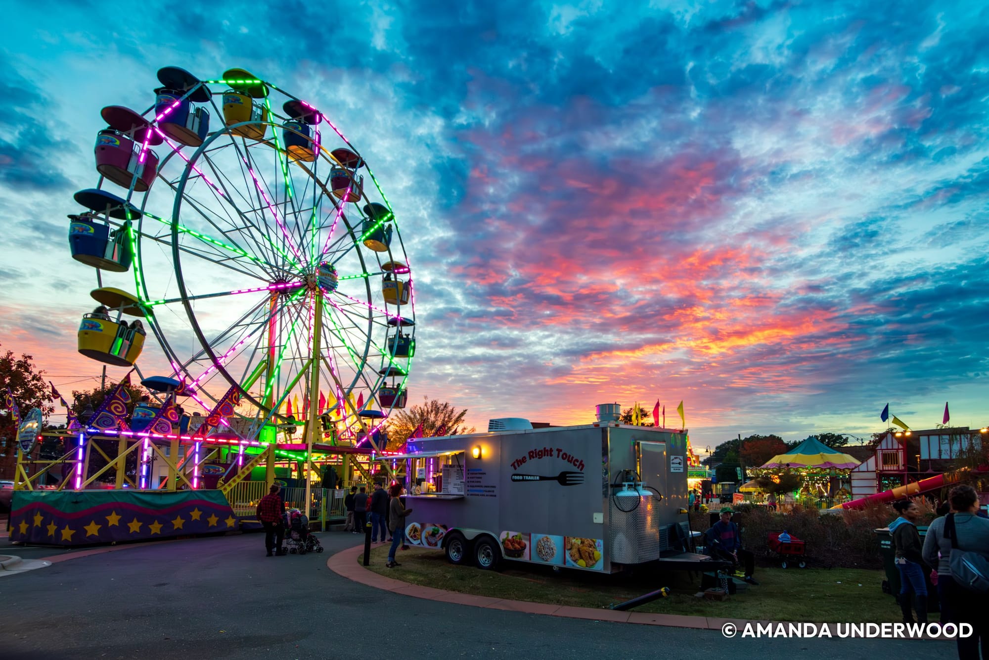 A brightly illuminated Ferris wheel rises above the Clayton Harvest Festival at sunset. Pink, orange, and blue clouds fill the sky as visitors walk among carnival rides and food trailers, including one labeled The Right Touch.
