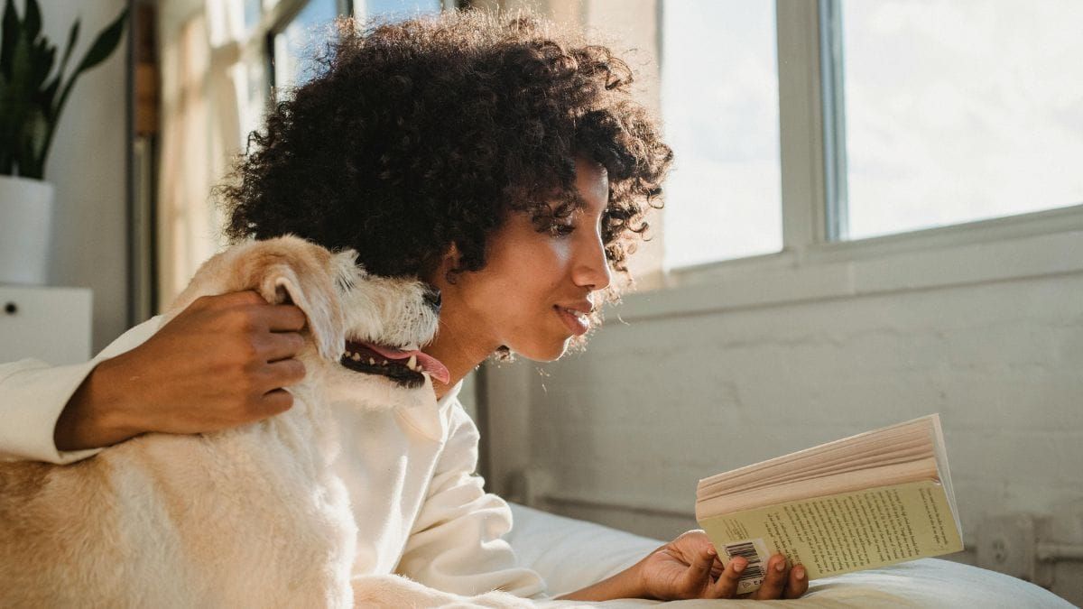 woman petting dog and reading book on bed