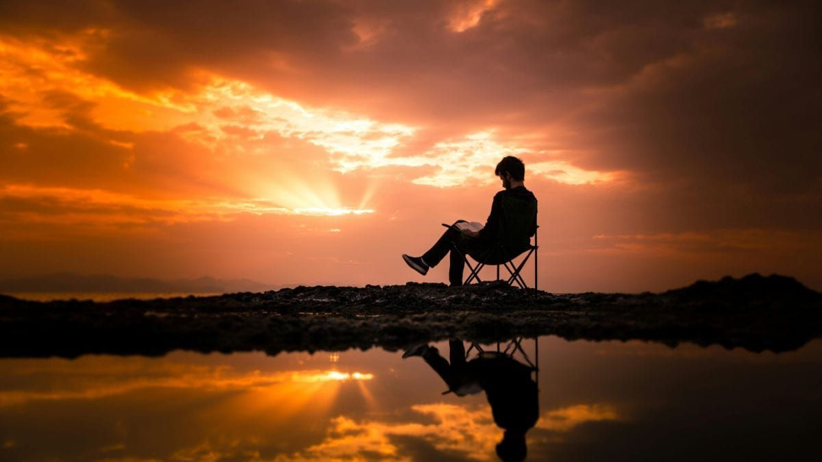 Person reading a book alone at sunset by the water with reflection in the foreground.
