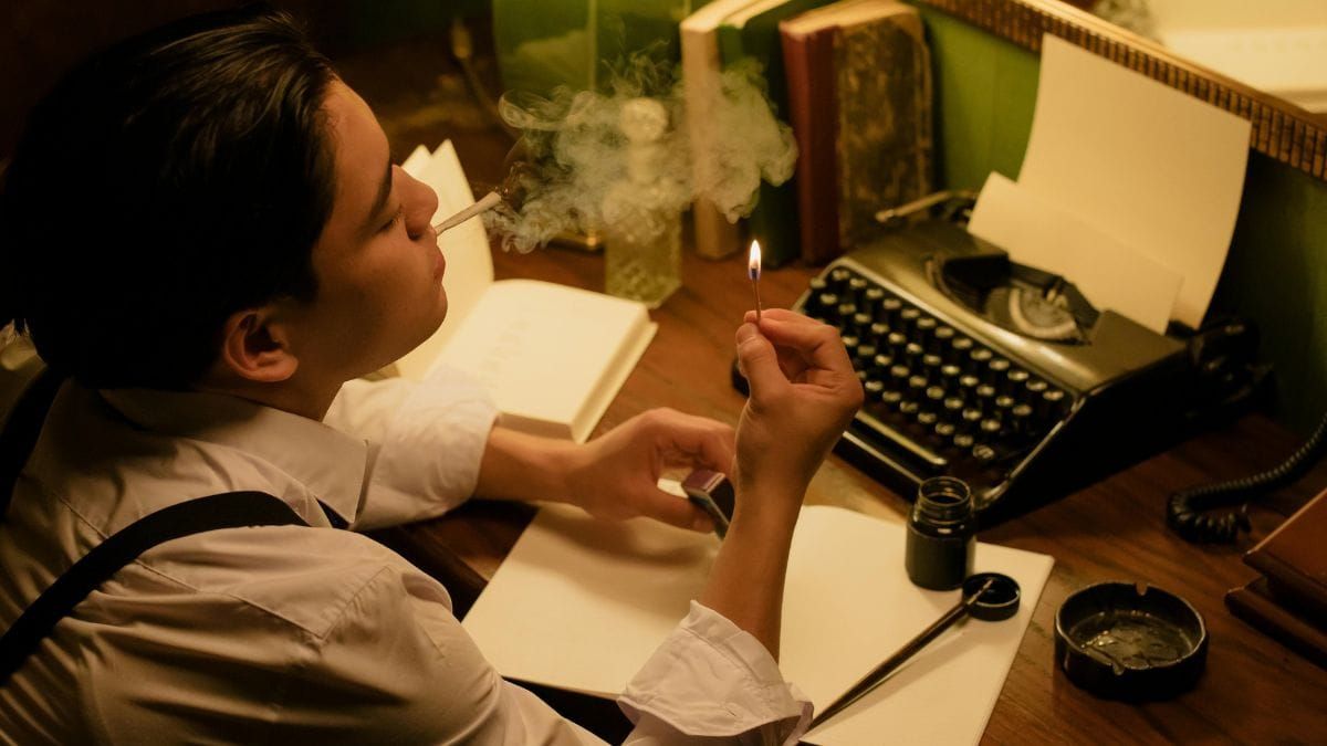 Person sitting at a desk writing beside a typewriter, lit by warm lamp light and cigarette smoke.