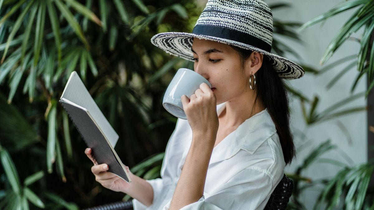 Woman reading a book and drinking coffee outdoors in a peaceful garden setting.