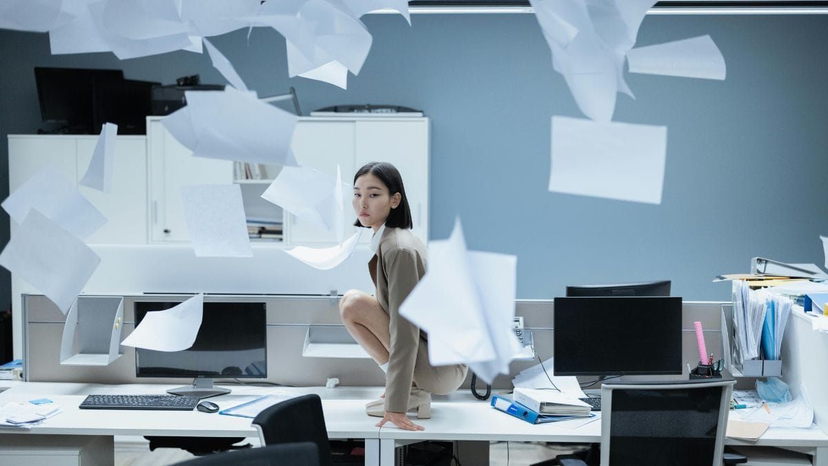 A woman crouches on a desk in a quiet office as papers float around her.