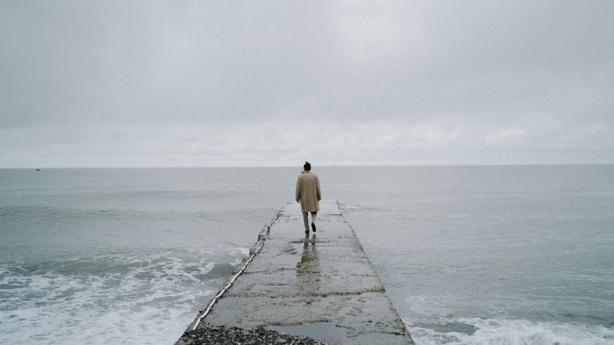 A person walking alone on a narrow pier beneath a grey sky above a calm sea.