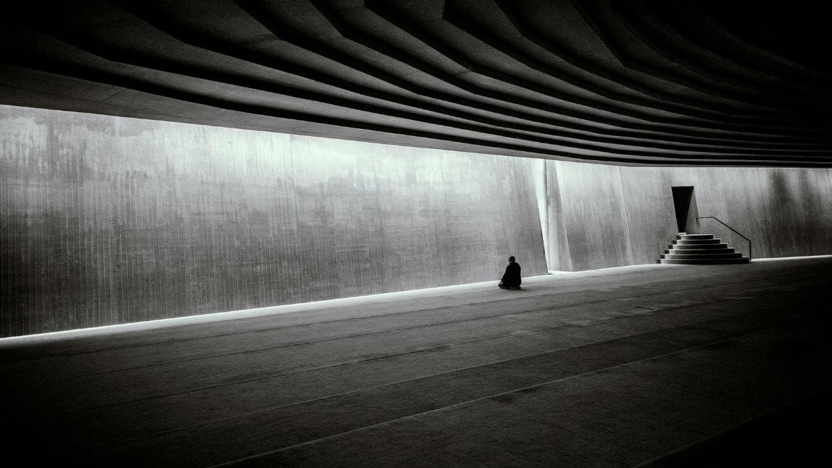 A solitary figure sits in a vast concrete space facing a illuminated wall, with stairs leading toward a dark exit.