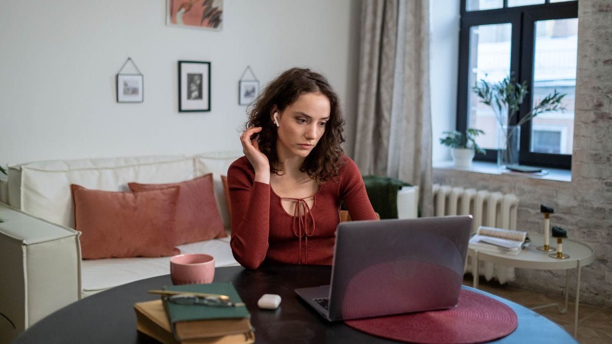 A woman working on a laptop at home with an earbud in.