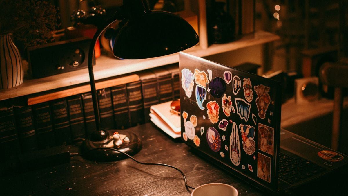 A sticker-covered laptop on a warm desk beside books and a lamp.