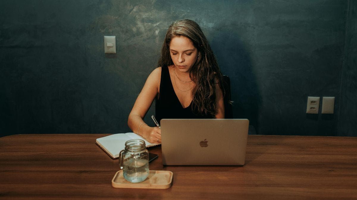 A woman writing in a notebook beside a laptop under a hanging lamp.