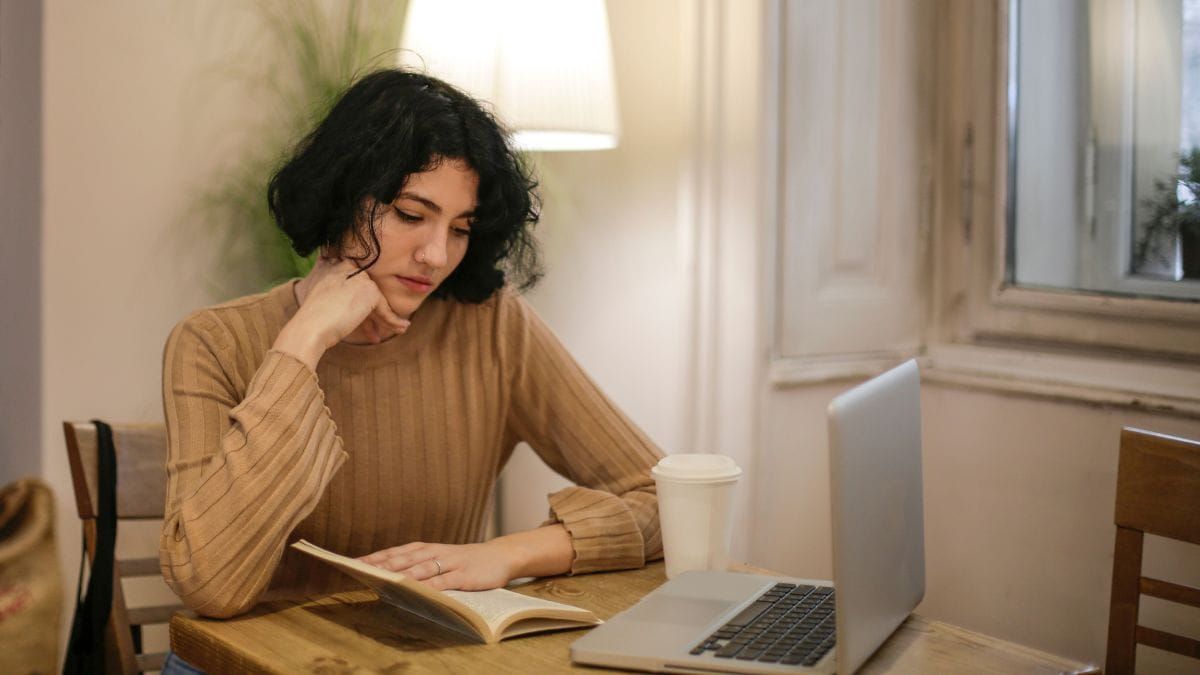 Person reading a book at a table with an open laptop nearby.