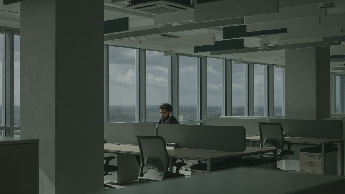 A lone office worker sitting among rows of empty desks in a modern open-plan office