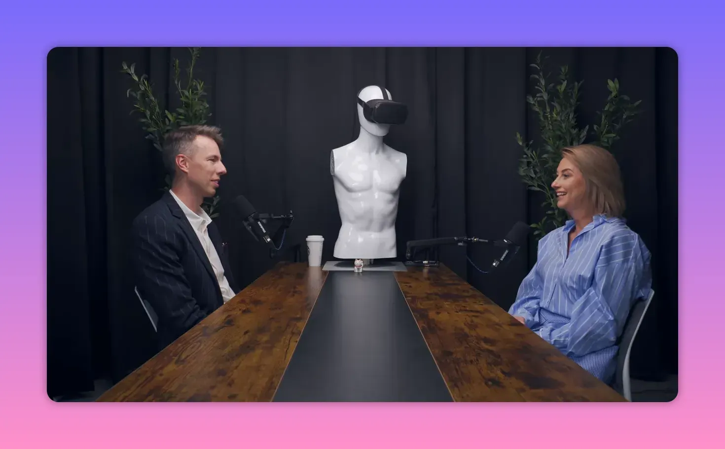 Широкий план интервью: ведущий и гость улыбаются за столом в студии, манекен в центре