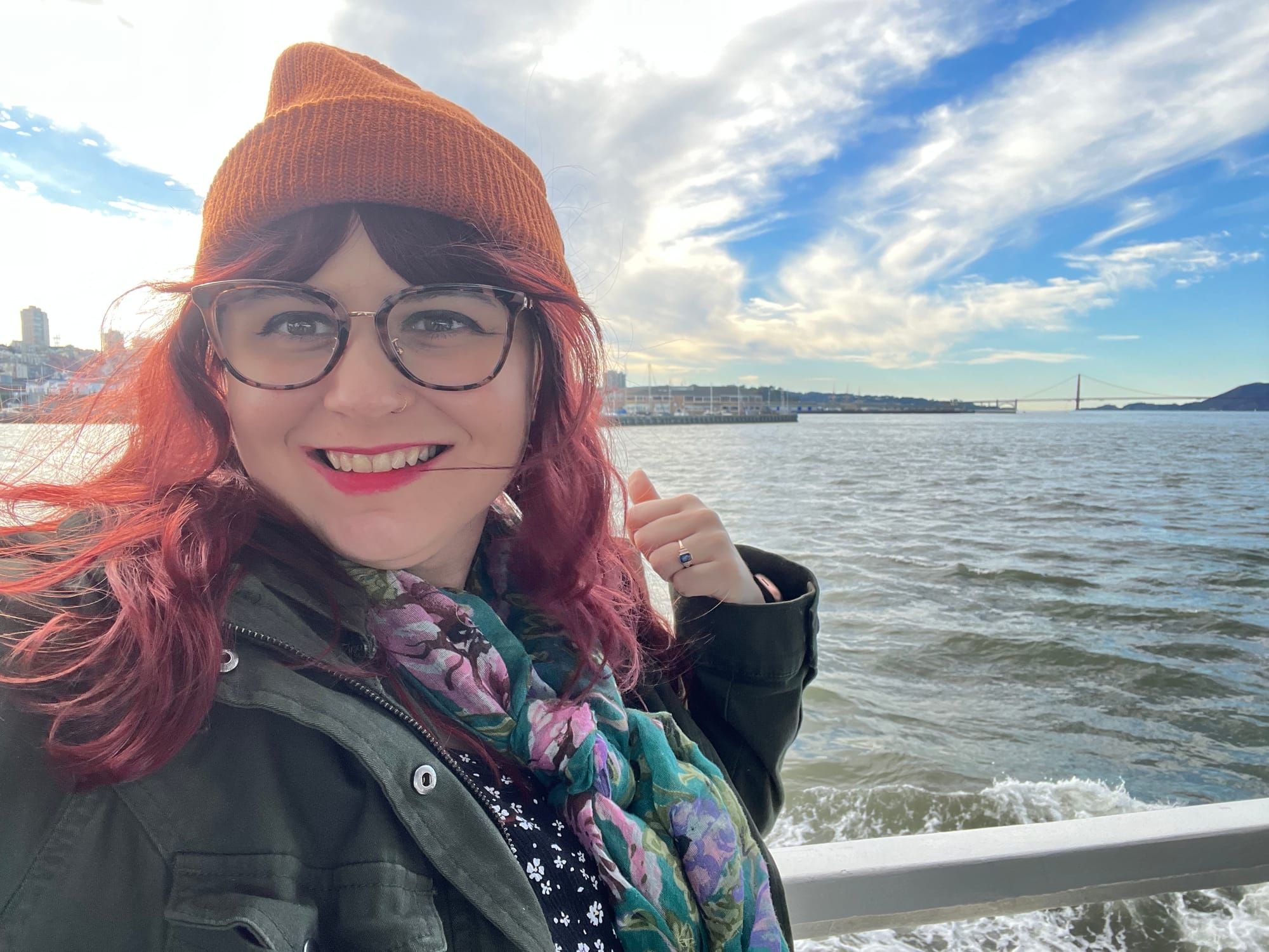 Cate Brecht, a white woman with red hair and glasses, poses at the railing of a ship in the San Francisco Bay. 