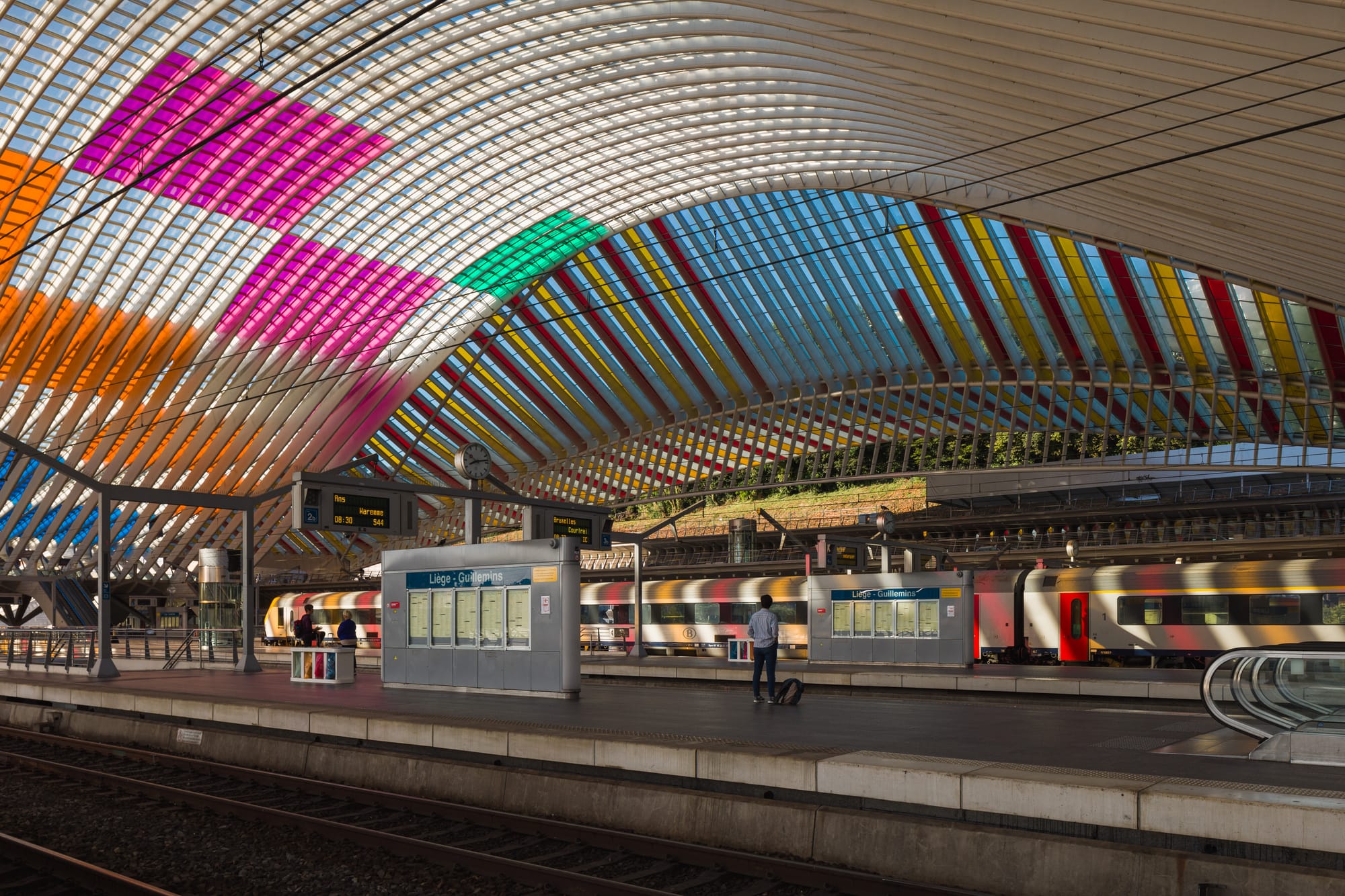 Interior of the Liège-Guillemins train station with people waiting for a train and colorful light spilling everywhere