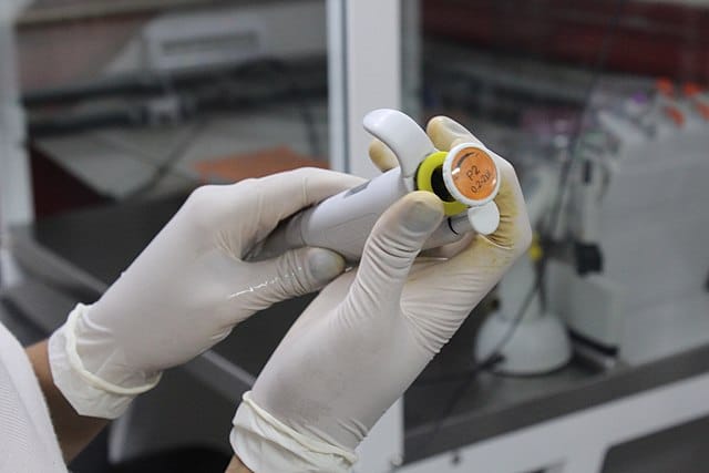 A pair of gloved hands holding a micropipette and twisting it to adjust it.