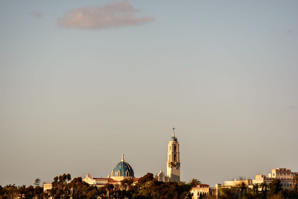 mostly a grayish sky with a solitary wispy cloud at the top of the frame, the bottom eighth of the image is a few buildings of the University of San Diego from a distance