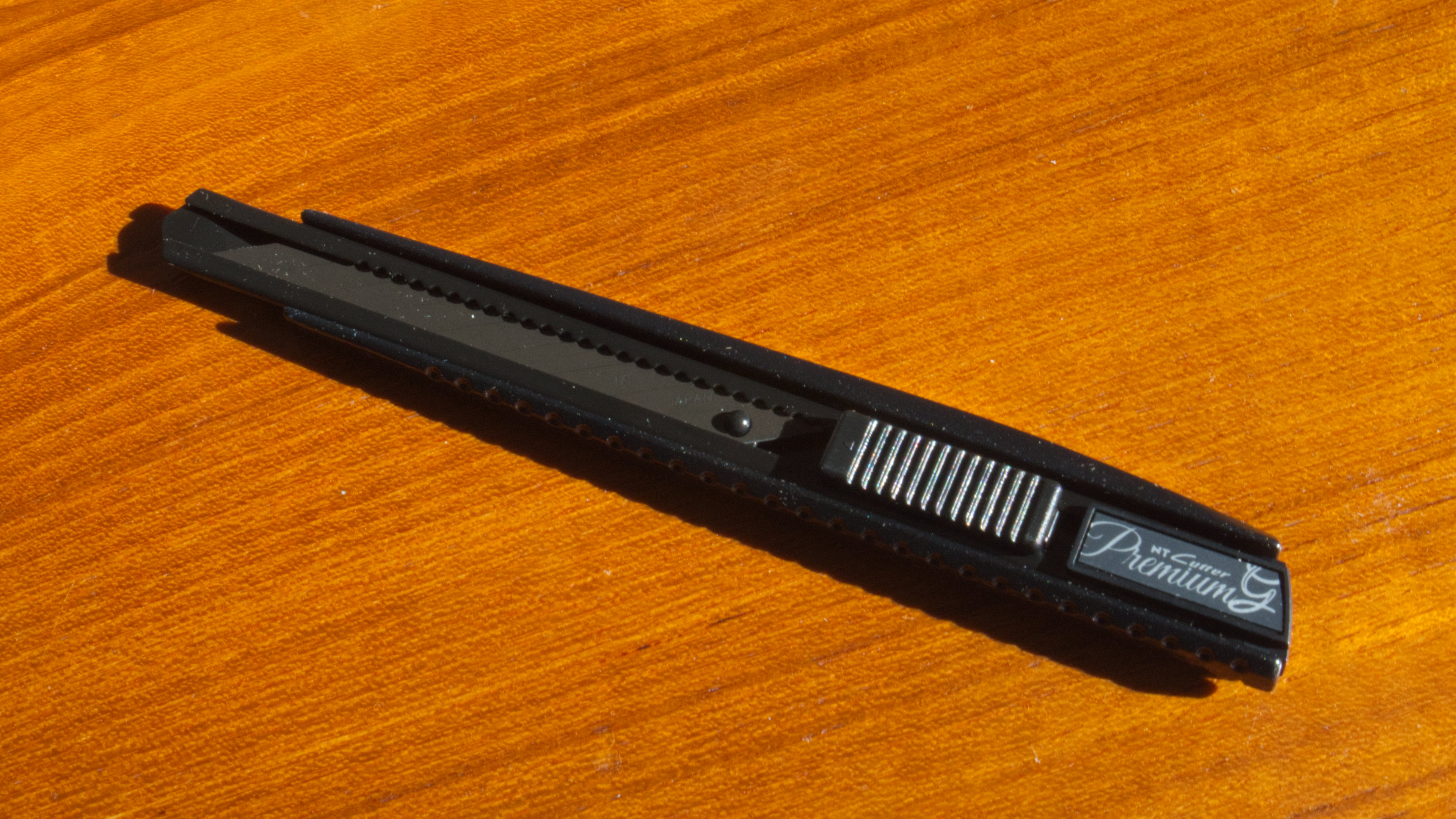 A black, metal NT Cutter Premium Evolution PMG series knife on a wooden backdrop.