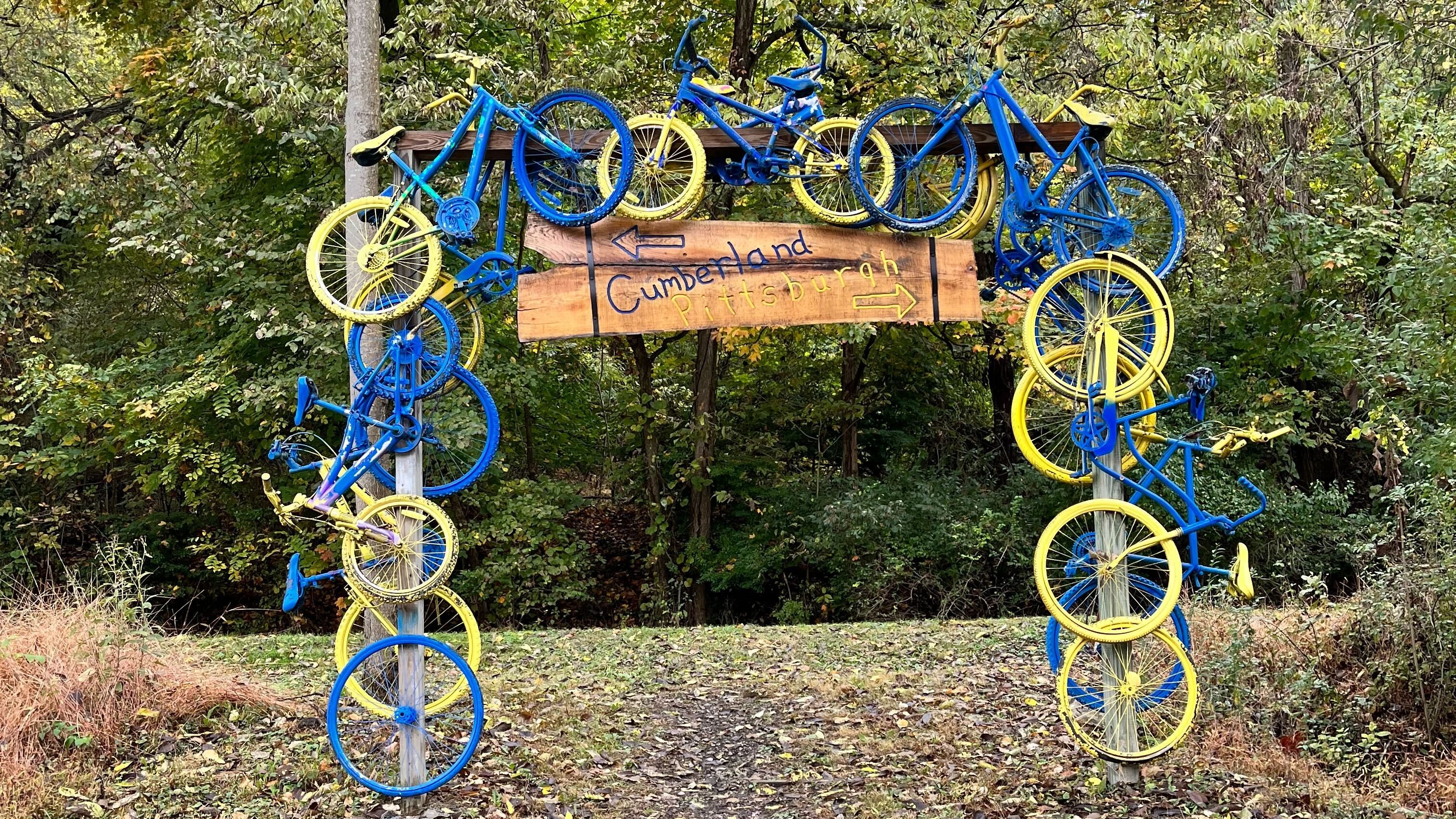 A wooden sign with blue and yellow bicycles attached. The sign text reads "Cumberland" and "Pittsburgh," with arrows pointing in opposite directions.
