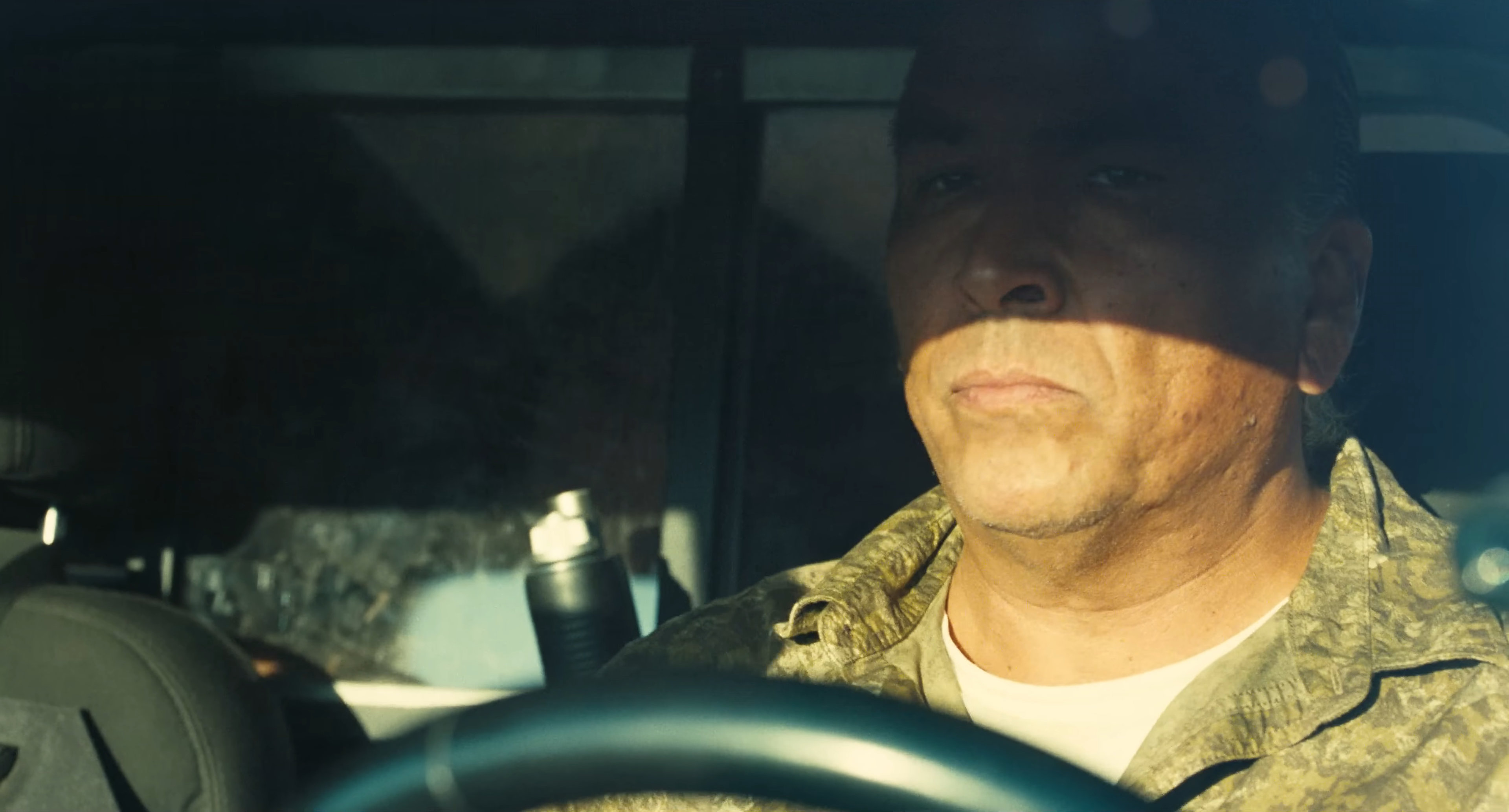 Eric Schweig as Avanti in One Battle After Another. He is driving a car, looking into the middle distance. There is a plexiglass barrier behind him.