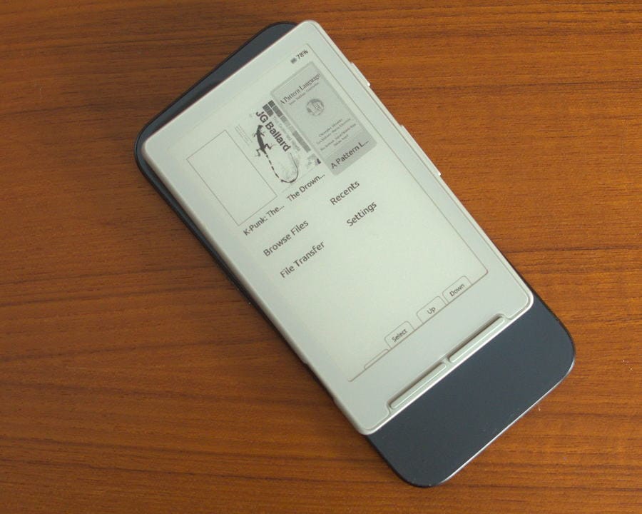 An Xteink on the back of a more recent iphone in a kind of metalic blue, the menu for CrossPoint is visible, you can see A Pattern Language, The Drowned World, K-Punk on the top of the recent list.