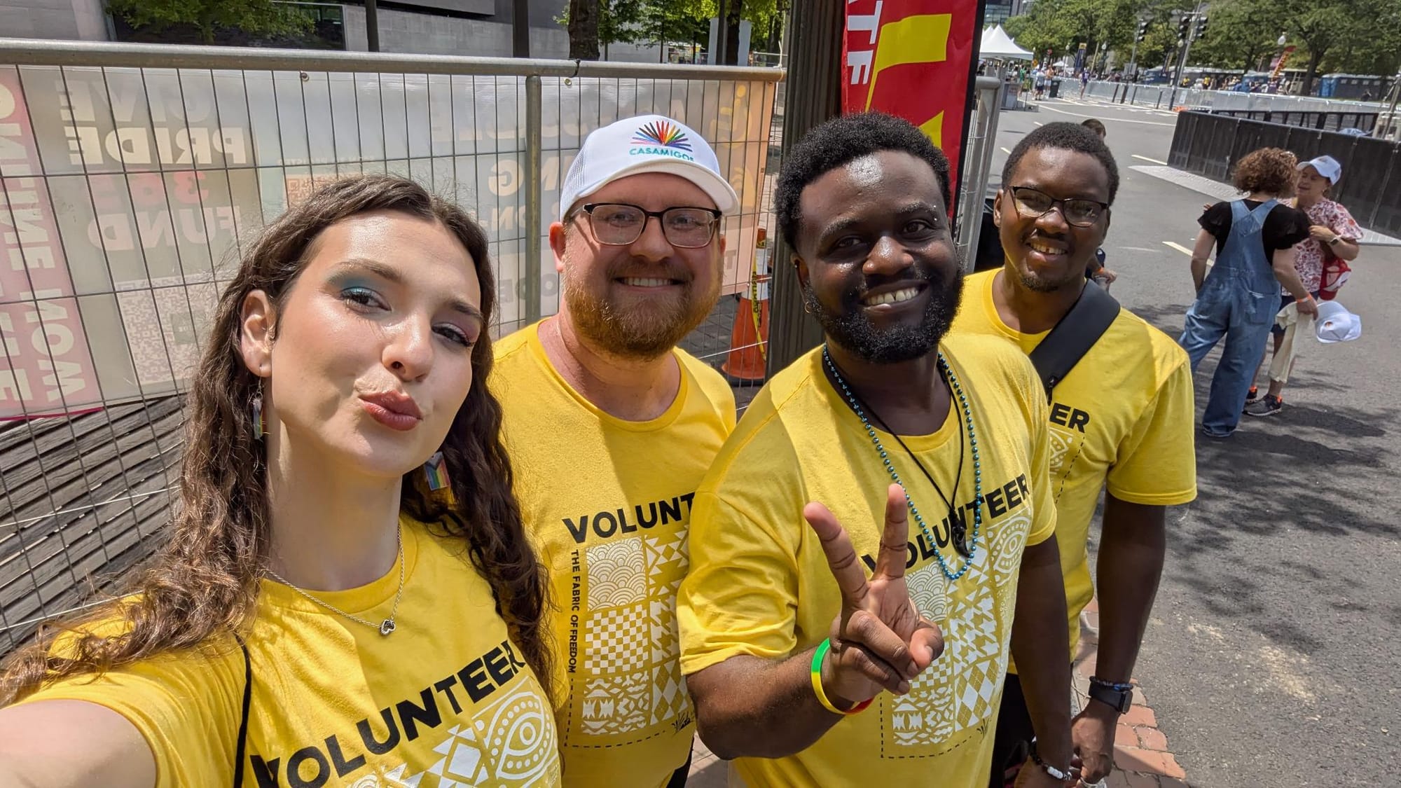 Photo depicting a group of four people, with Ludovic Mbock an African man second from the right giving a peace sign while wearing a yellow shirt that reads 'volunteer'.