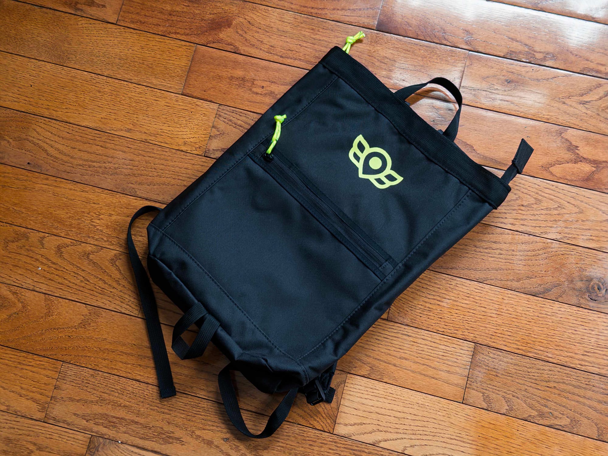 A backpack is on a wooden floor. There are yellow zipper pulls, and the bike angels logo in bright yellow.