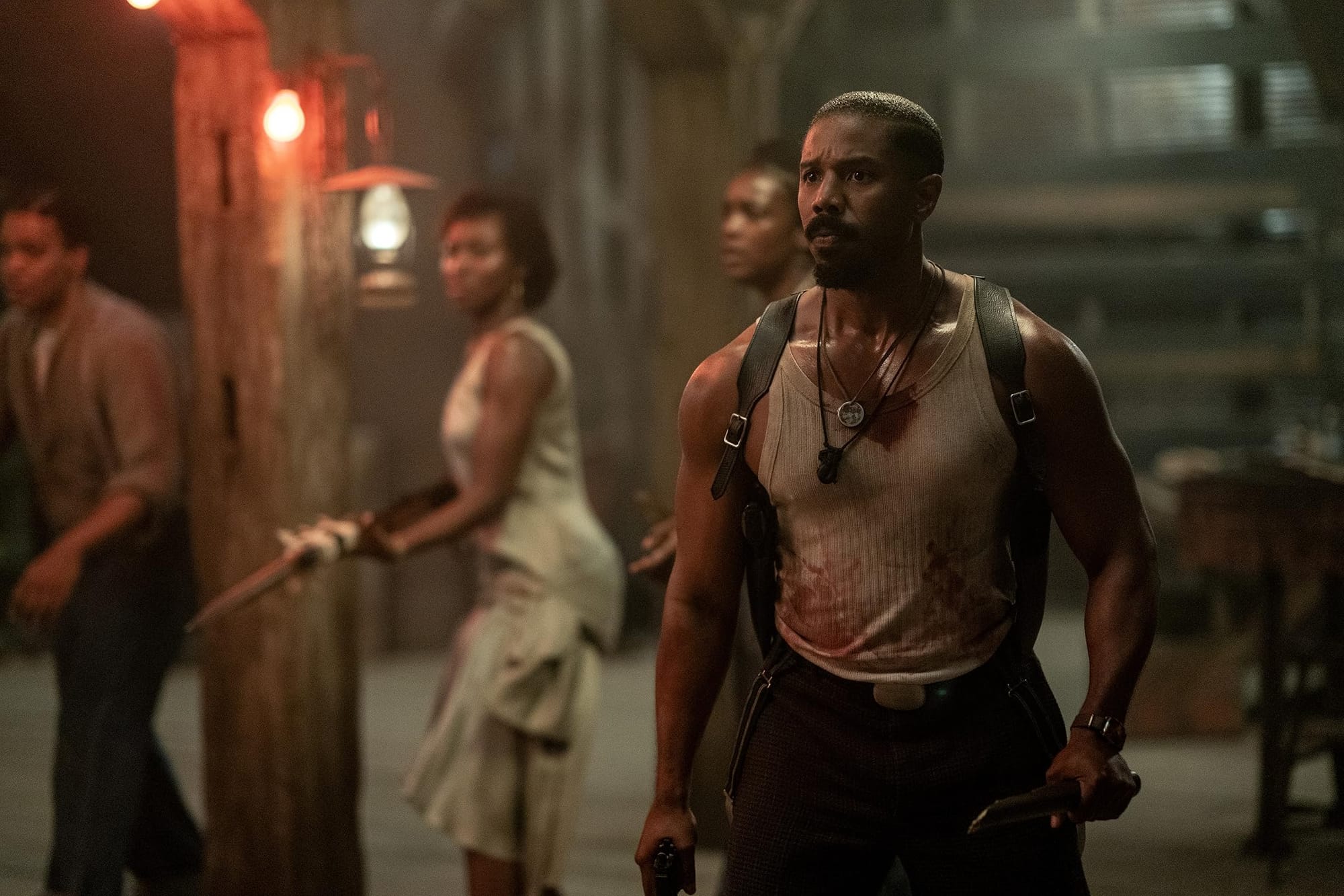 Michael B. Jordan as Stack, in a bloodied tank top and shoulder holsters holding a knife at his side while others brace for attack out of frame.