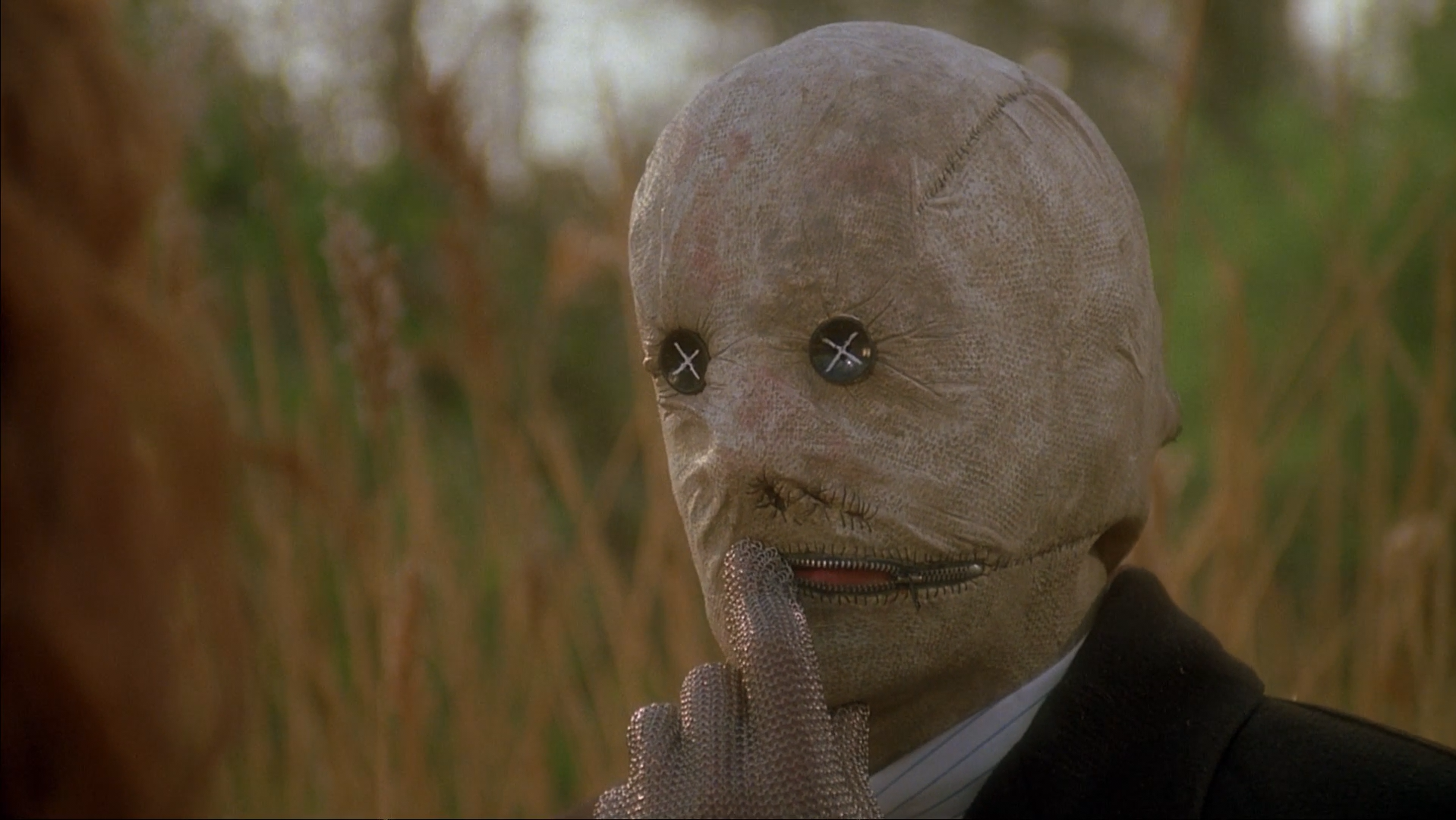 A man in a suit and a scary mask with buttons for eyes and a zipper mouth, his hand is gloved in chainmail.