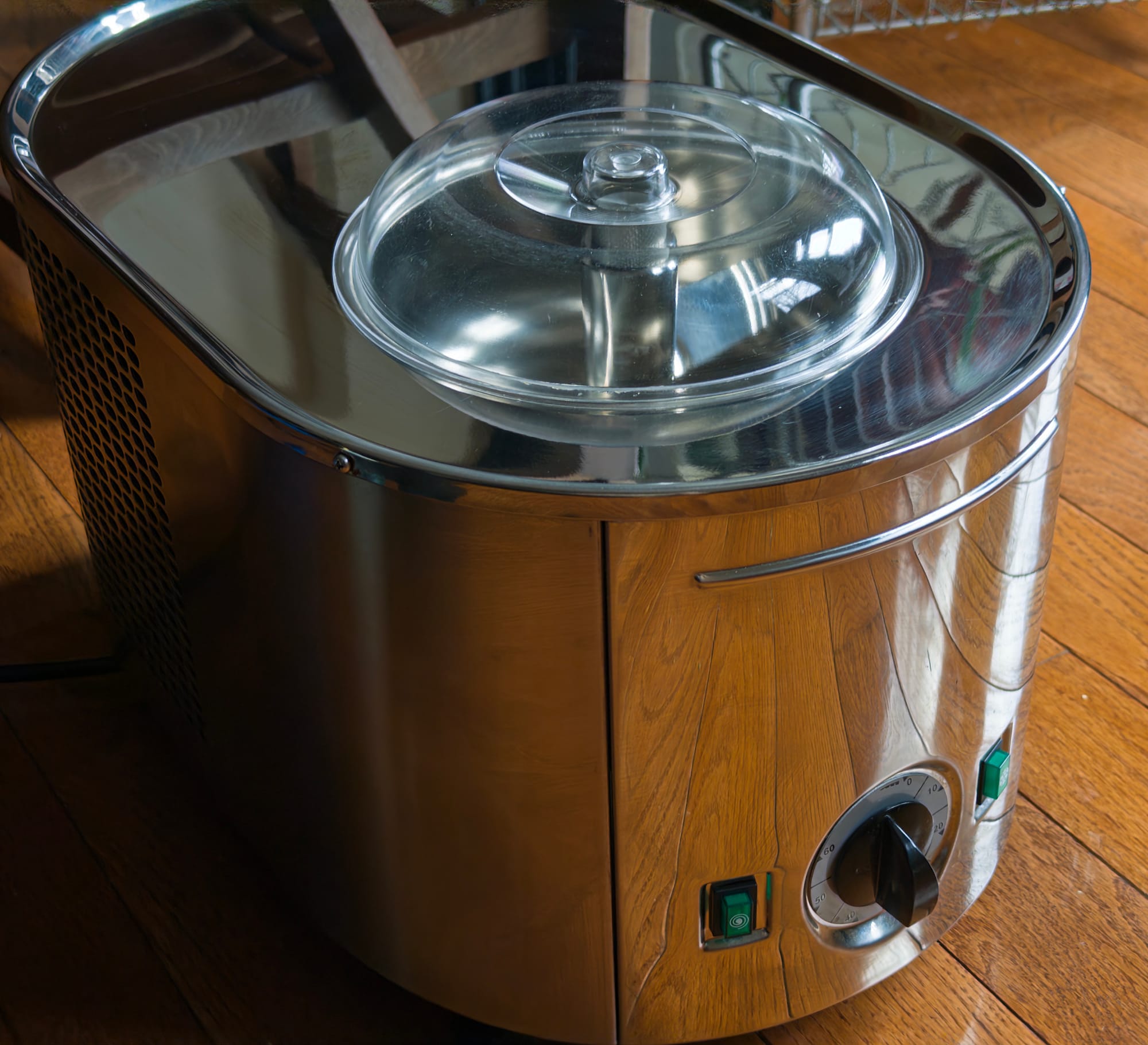 A beautiful silver ice cream machine with tactile buttons and dials. It looks like a sexy italian sports car from the 80s. The chrome finish reflects the wood, and there is a plastic dome on top.