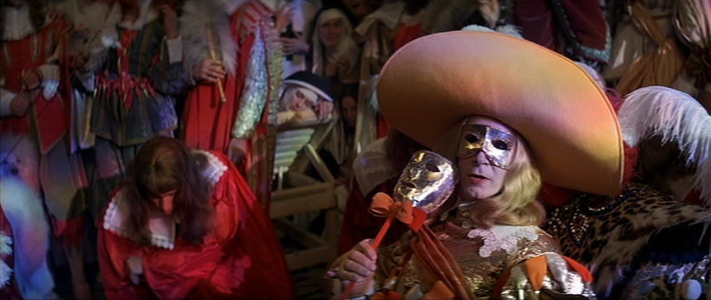 A foppish nonbleman lit by rainbow gels with a giant hat, a mask, and holding a hand mask. Nuns and various people are in the background.