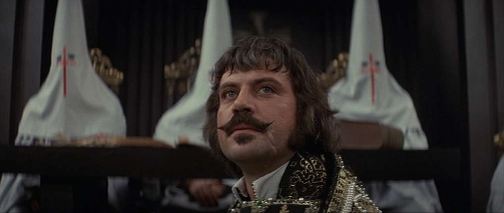 Oliver Reed looking defiantly as robed inquisitors are in the background out of focus.