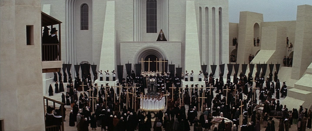 A large set of a town's square with towering buildings, a crowd in black, and banners.