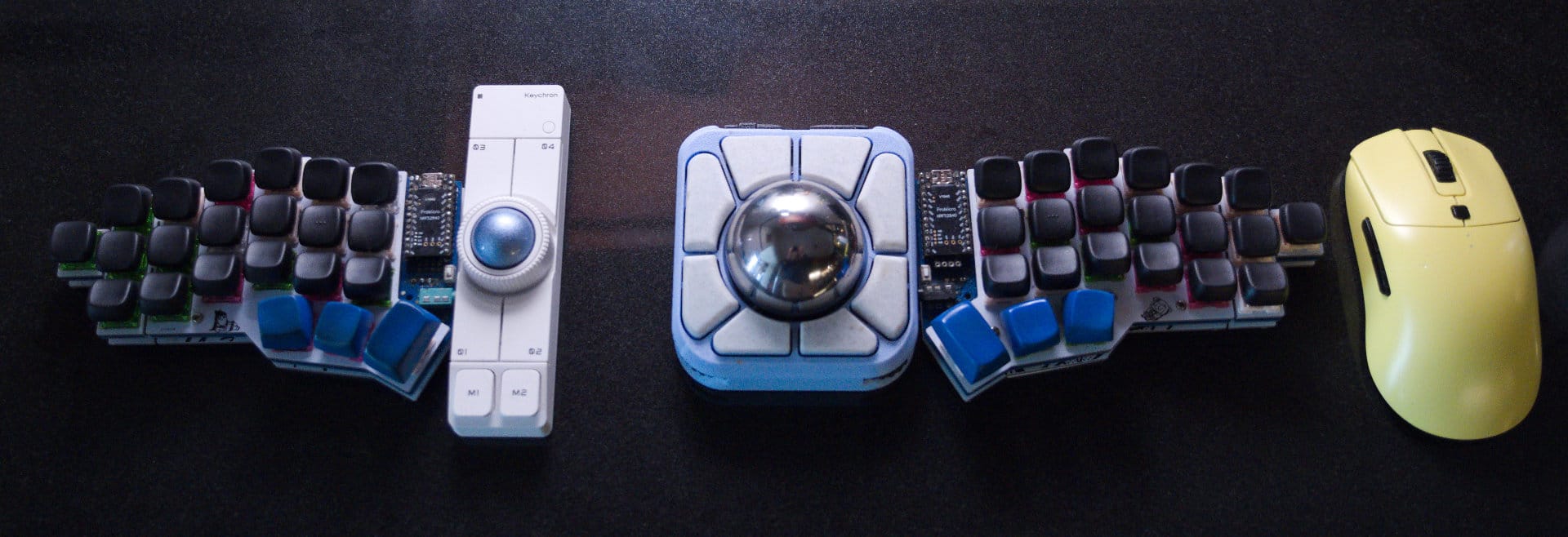 Two sides of a split keyboard and two trackballs in between. One is large and blue with white buttons and a steel ball. The left one is the nape pro. A yellow Vaxee mouse is on the right.