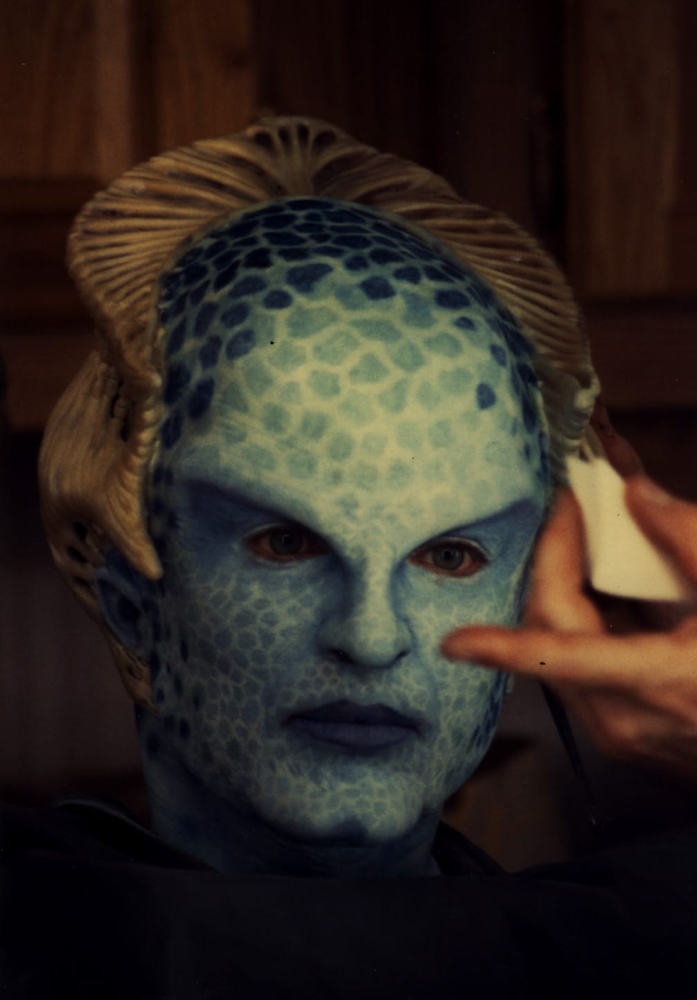 Actress Mira Furlan in a sea green skin tone make up that sprinkled with large dark blue freckles and wearing a gold bone crown. 