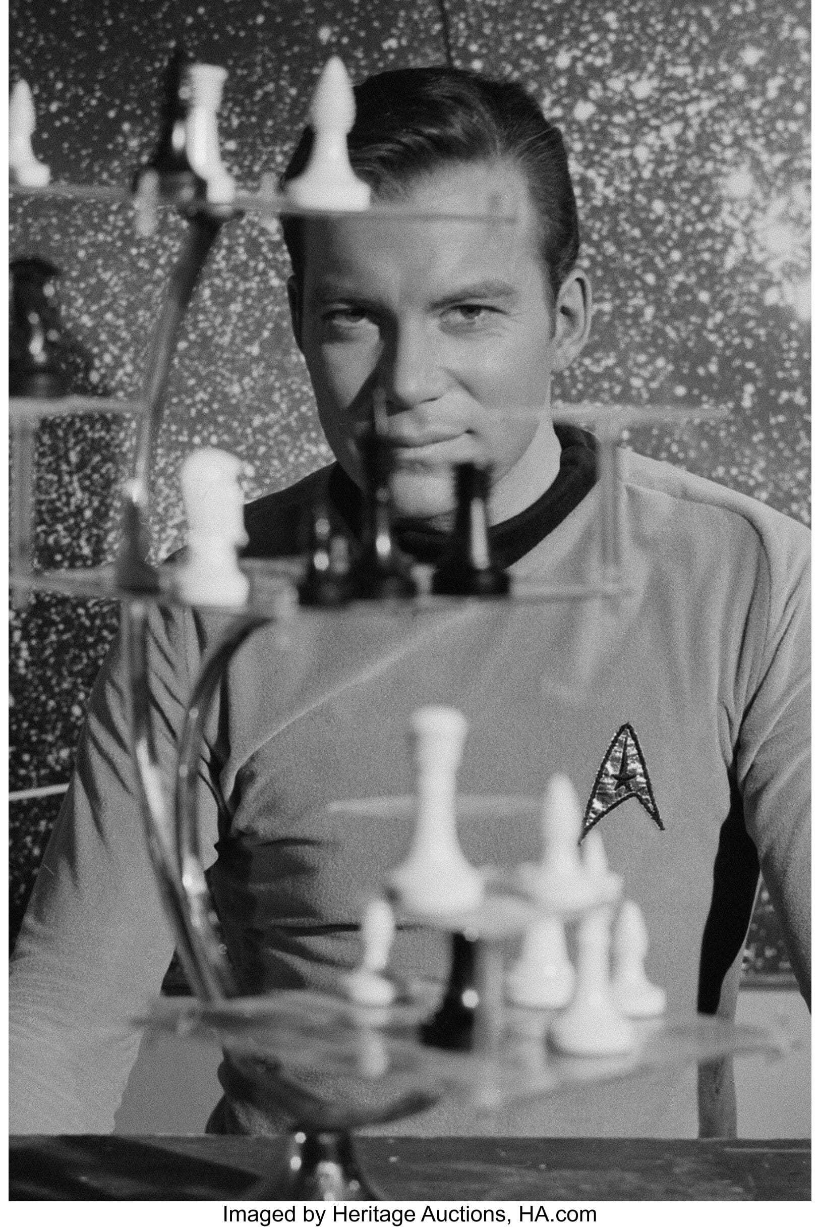 Captain James T. Kirk sits behind a three-dimensional chess set, blurred in foreground. Behind Kirk is a starmap. 