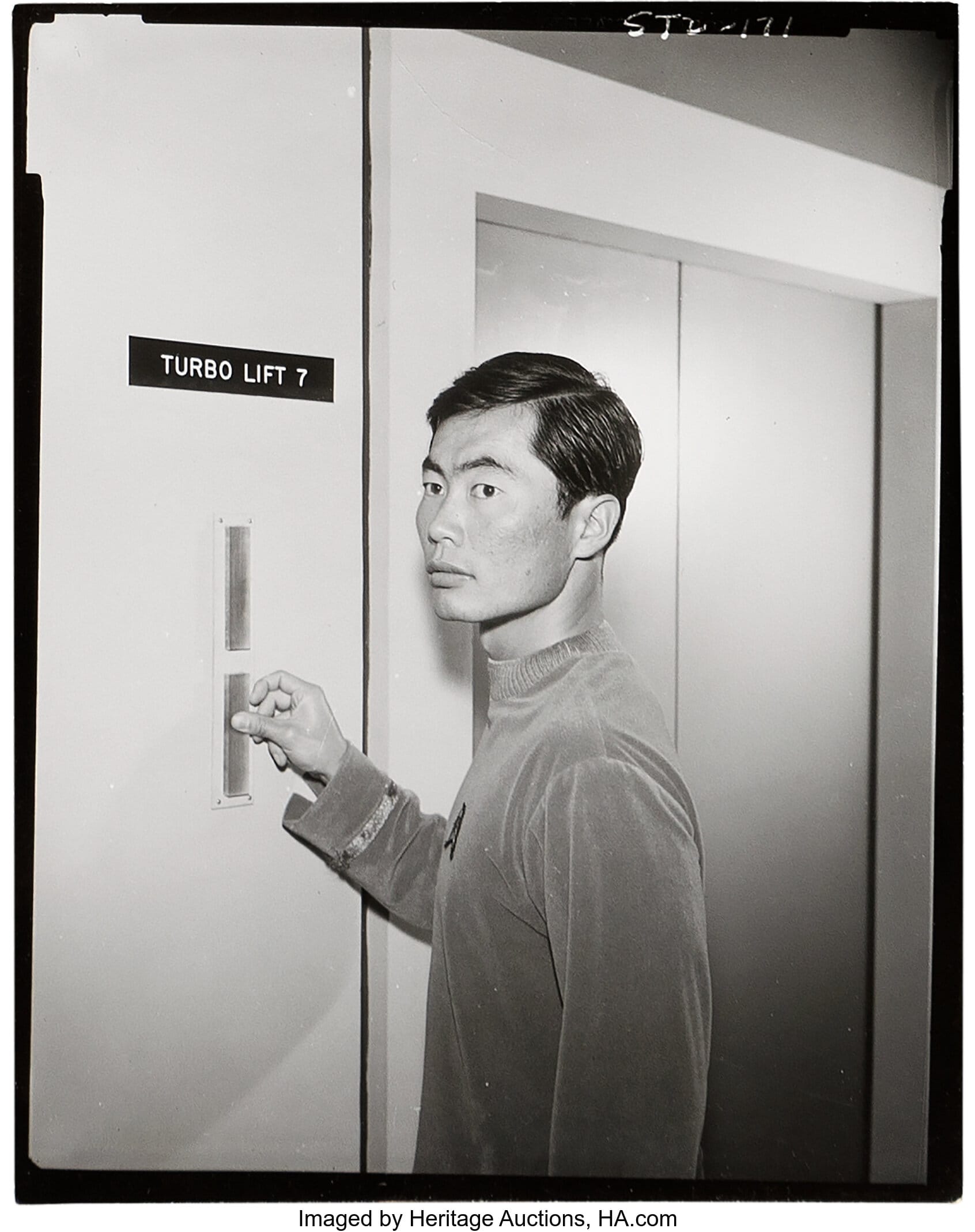 Lt. Sulu in a B&W picture from the second STAR TREK pilot. He is in a blue ribbed pilot uniform, pressing the turbolift call button as he stands next to the lift doors. A sign above the call buttons reads: Turbo Lift 7.  