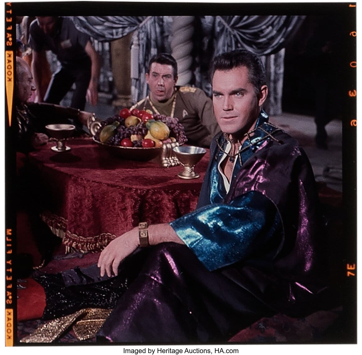 Jeffrey Hunter as Captain Pike in sparkly Arabian night robes sitting on pillows. He sits at a round table with gold goblets and a bowl of fruit. It's draped in a red table cloth with gold tassels. A Starfleet officer sits at the other end of the table.  