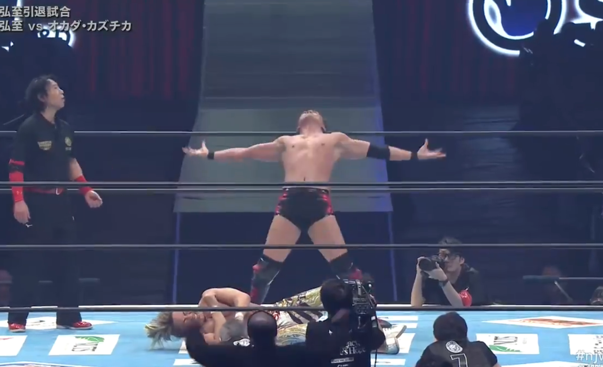 Okada strikes the Rainmaker Pose, both arms stretched out, palms up.